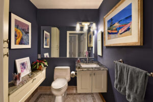 Modern bathroom with dark walls, framed art, a toilet, sink with granite countertop, and gray towel hanging on the wall.